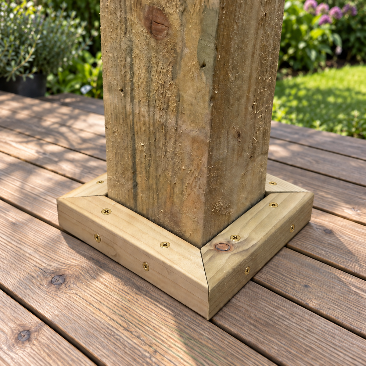 A square wooden 150 x 150mm post base fitted with four corner brackets and secured with screws, shown installed on a wooden deck with a garden background.