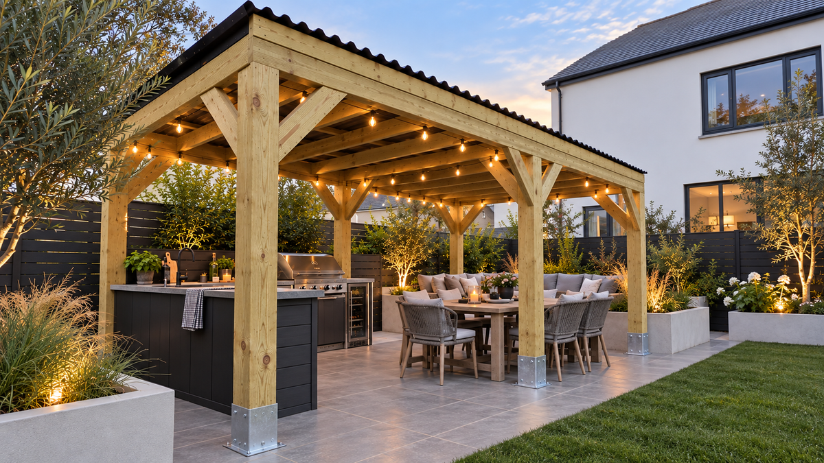 A large wooden pergola featuring a black roof with an integrated dark outdoor kitchen island and dining area, set in a modern back garden with festoon string lights on at dusk.