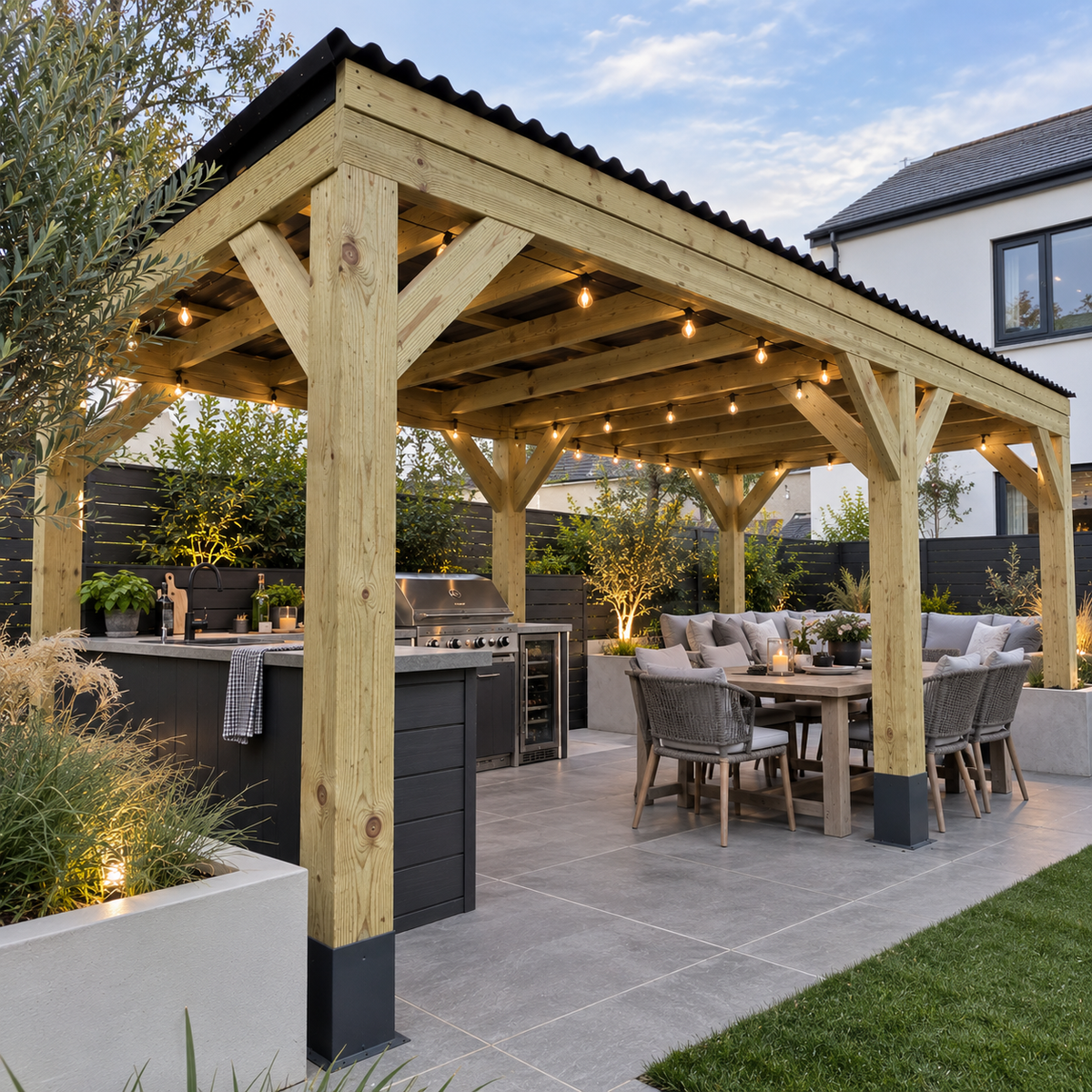 A large outdoor wooden pergola with integrated outdoor kitchen island and string lights, shown in a landscaped backyard setting with a dining table, patio seating, plants, and a house in the background.