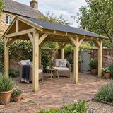 A large heavy duty wooden garden gazebo with a dark felt shingle roof in a brick patio backyard, featuring a wicker armchair with a navy throw, a small round wooden table, and a garden bench under the gazebo.