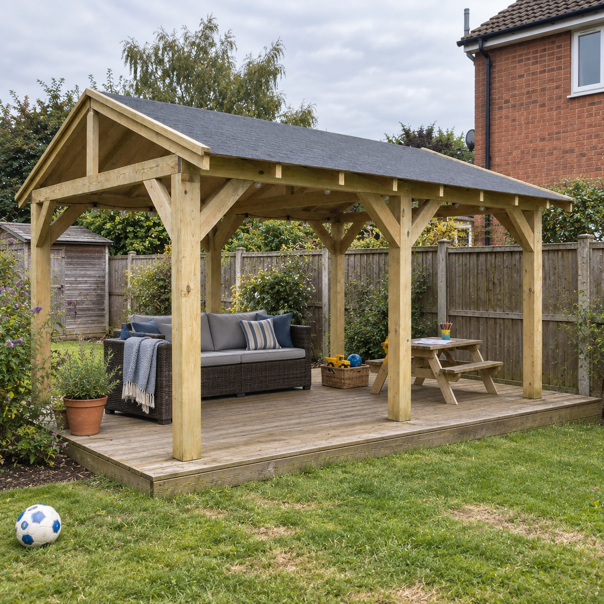 A large heavy duty wooden garden deck with a dark grey felt roof, situated on a raised wooden deck in a UK back garden. The wooden decking is furnished with a dark grey wicker sofa, a small wooden picnic table, and a throw blanket.