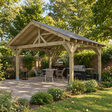 A large wooden garden gazebo with a dark felt shingle roof is shown in a UK landscaped back patio garden setting, featuring a dining table and chairs, a grill, and outdoor greenery.