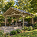 A large wooden garden gazebo with a dark felt shingle roof is shown in a UK landscaped back patio garden setting, featuring a dining table and chairs, a grill, and outdoor greenery.