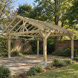 A large wooden gazebo frame with chunky posts  and a pitched roof and open side panels, shown installed on a paved patio in a landscaped UK garden with trees, shrubs, and a brick wall.