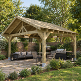 A large heavy duty wooden gazebo with a pitched roof is shown in a UK landscaped back garden. Under the cover, there is a stone patio area with a gray sofa, dining chairs, lanterns, a grill, and a small kitchen island.