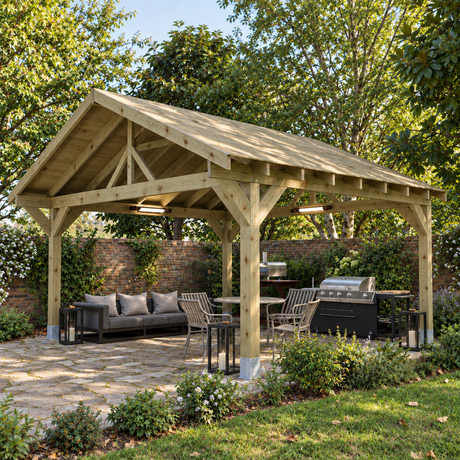A large heavy duty wooden gazebo with a pitched roof is shown in a UK landscaped back garden. Under the cover, there is a stone patio area with a gray sofa, dining chairs, lanterns, a grill, and a small kitchen island.