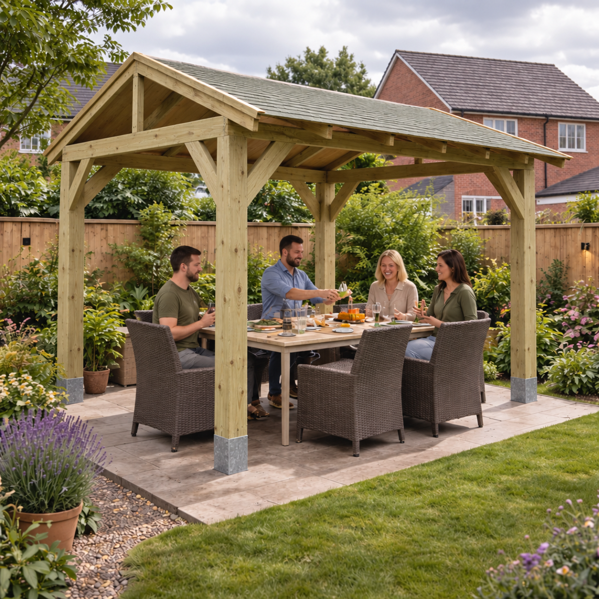 A Timbakit wooden garden gazebo with a waterproof shingle roof, a dining table and four woven patio chairs, with four people sitting underneath and drinking. The gazebo is set on a paved patio in a landscaped garden with grass, plants, and a wooden fence, with a brick house visible in the background.