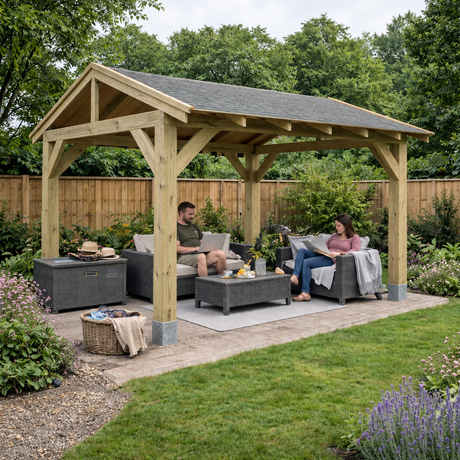 A large wooden garden gazebo with a grey felt shingle roof in outdoor seating area with two people sitting on grey sofas and a coffee table. The scene is set in a landscaped garden with a wooden fence, green trees, and a paved patio.
