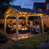 A wooden outdoor patio garden gazebo with warm festoon lights is set in a backyard at dusk, with four people sitting underneath on patio furniture and a coffee table.