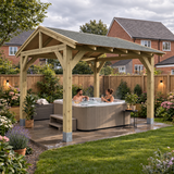A large wooden gazebo-style patio cover with a grey felt shingle roof over a hot tub, with two people sitting inside in a landscaped back garden. The scene includes grass, flowers, a wooden fence, and neighboring houses under a cloudy sky.