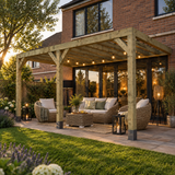 A large wooden lean to box pergola over a paved patio with a wicker sofa, armchairs, coffee table, string lights, and outdoor lanterns, set in a landscaped garden in front of a brick house at dusk.