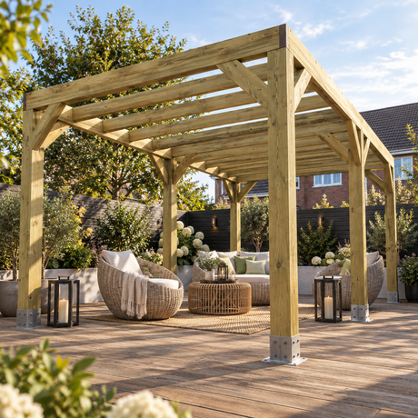 A large wooden box pergola with a natural light wood finish stands on a wooden deck in a landscaped backyard. Underneath the pergola is a seating area with wicker armchairs, a round coffee table, and a sofa with cushions.