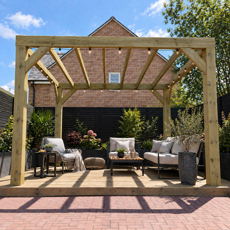 A chunky wooden box pergola with integrated string lights stands over a raised patio deck furnished with a sofa, armchair, coffee table, and potted plants. A brick house and trees are visible in the background under a blue sky.