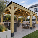 A large outdoor wooden pergola with integrated outdoor kitchen island and string lights, shown in a landscaped backyard setting with a dining table, patio seating, plants, and a house in the background.