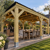 A large wooden outdoor patio pergola with a black roof, shown in a landscaped backyard. Underneath, a wooden dining table and wicker chairs are arranged with string lights and pendant lamps.