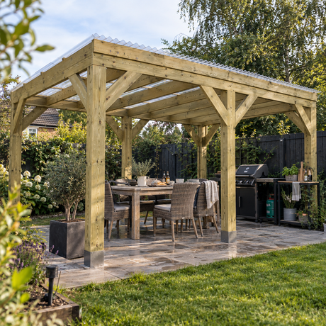 A large wooden outdoor patio pergola with a corrugated clear roof, set in a landscaped uk back garden with a dining table, chairs, a grill, and green grass.